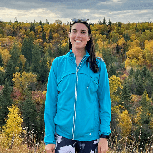 Laura in a blue sweater smiling at the camera over looking a forest in fall.